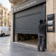 Instalador junto a una persiana metu00e1lica gris a medida y muestras de perfiles en la entrada de un garaje en Barcelona.