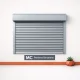 Persiana metálica comercial gris centrada sobre fondo blanco, con maceta de suculenta y base terracota, minimalista y serena. Cuales son las persianas metalicas mas recomendadas para locales comerciales en Barcelona.