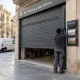 Instalador junto a una persiana metu00e1lica gris a medida y muestras de perfiles en la entrada de un garaje en Barcelona.